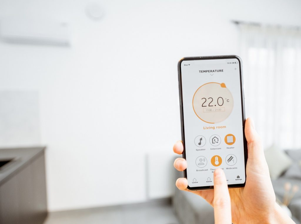 En hånd holder en mobiltelefon opp og vi ser en app som styrer varmen fra telefonen. En pekefinger trykker på termostatknappen på telefonen